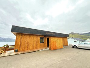 A wooden cabin with a black roof, labeled 'RINKUHUSINI,' situated by a body of water with mountains in the background.