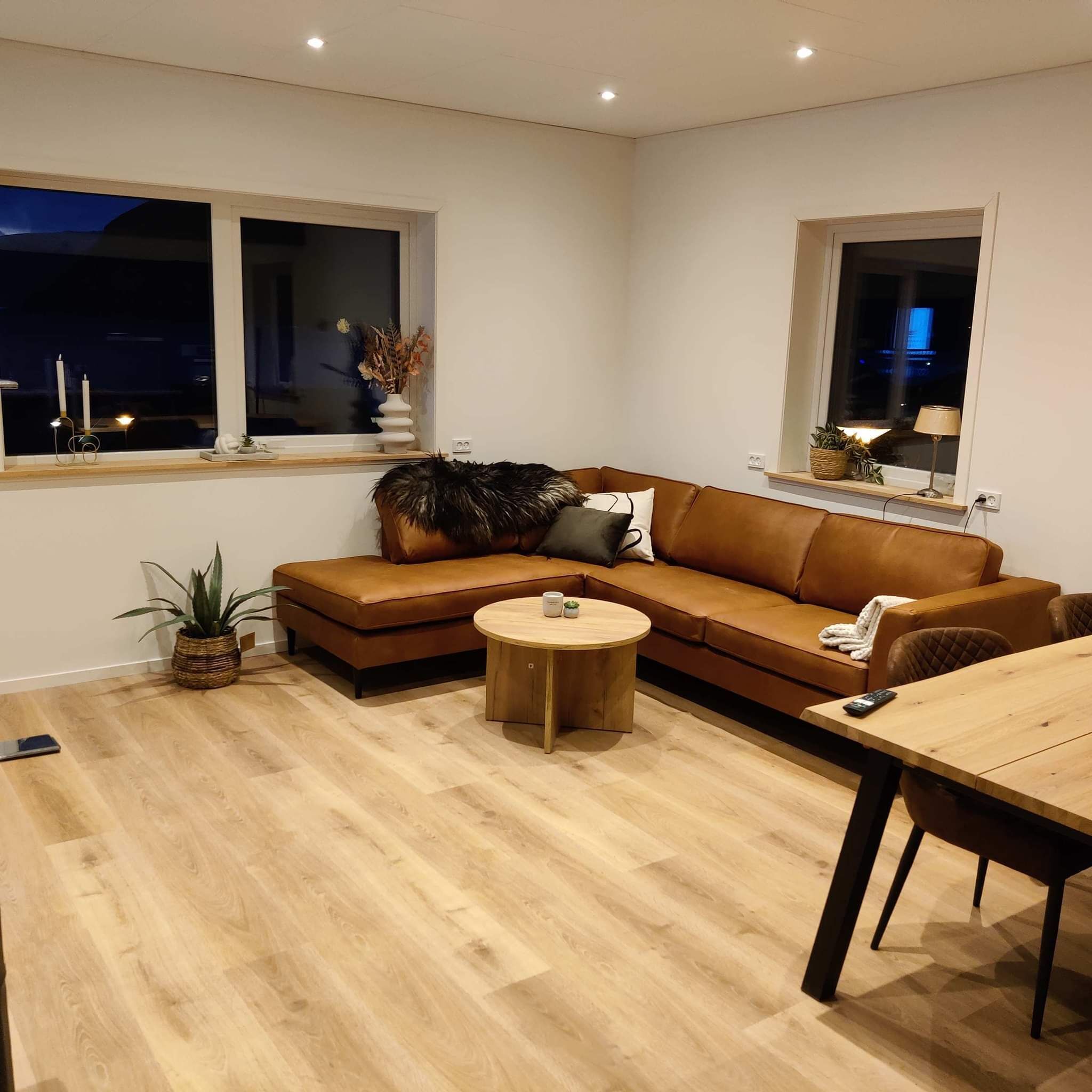 Ein modernes Wohnzimmer mit einem braunen Ledersofa, einem runden Couchtisch und einem Esstisch mit Stühlen. Im Raum steht eine Topfpflanze und auf der Fensterbank eine Vase mit Blumen.