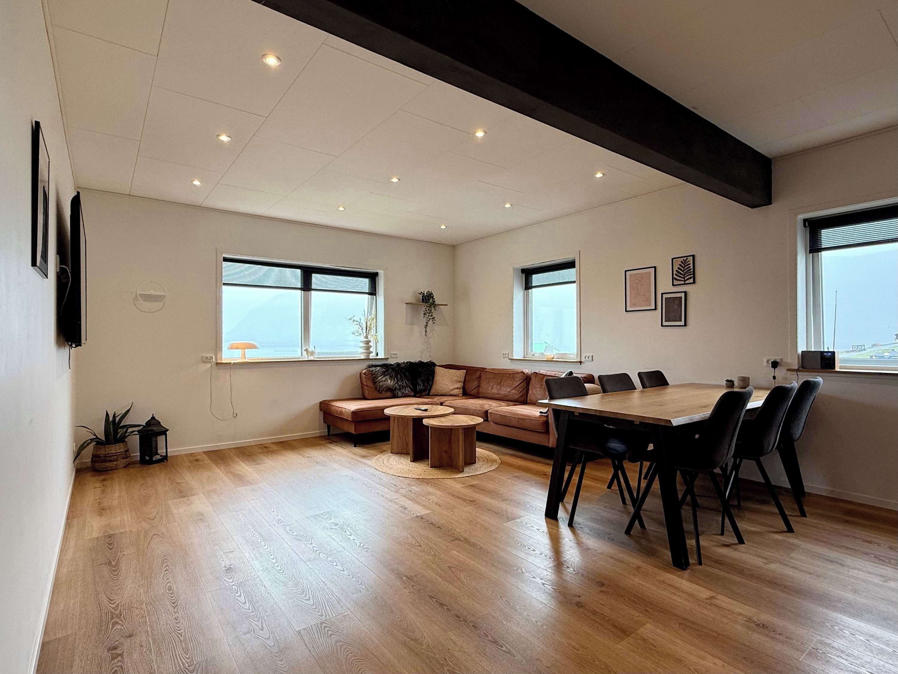 A spacious living area with a wooden floor, a large sofa, a dining table with chairs, and multiple windows providing natural light.