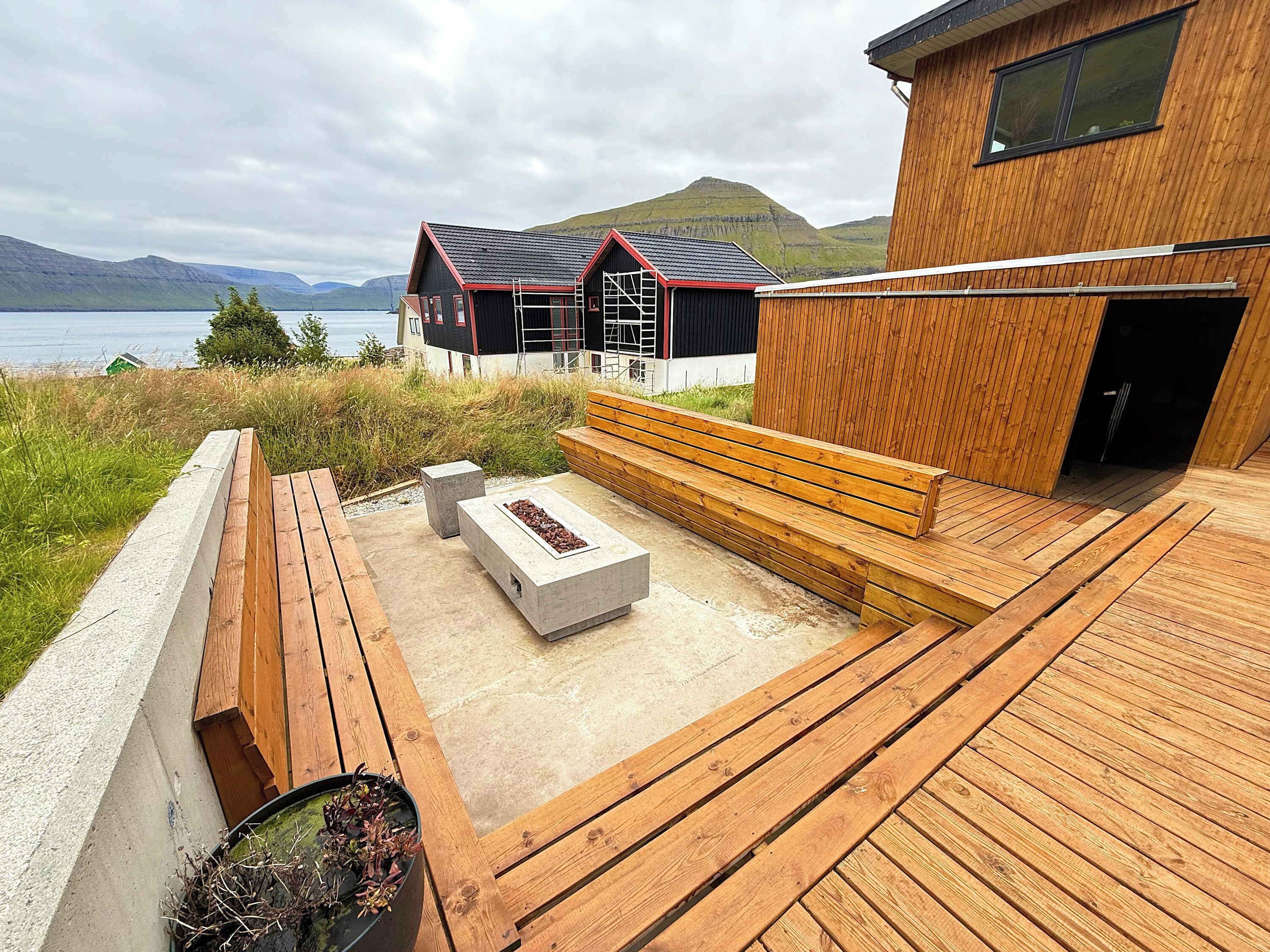 Eine Holzterrasse mit Sitzgelegenheiten und einem Feuerkorb, mit Blick auf ein Gewässer und Hügel.