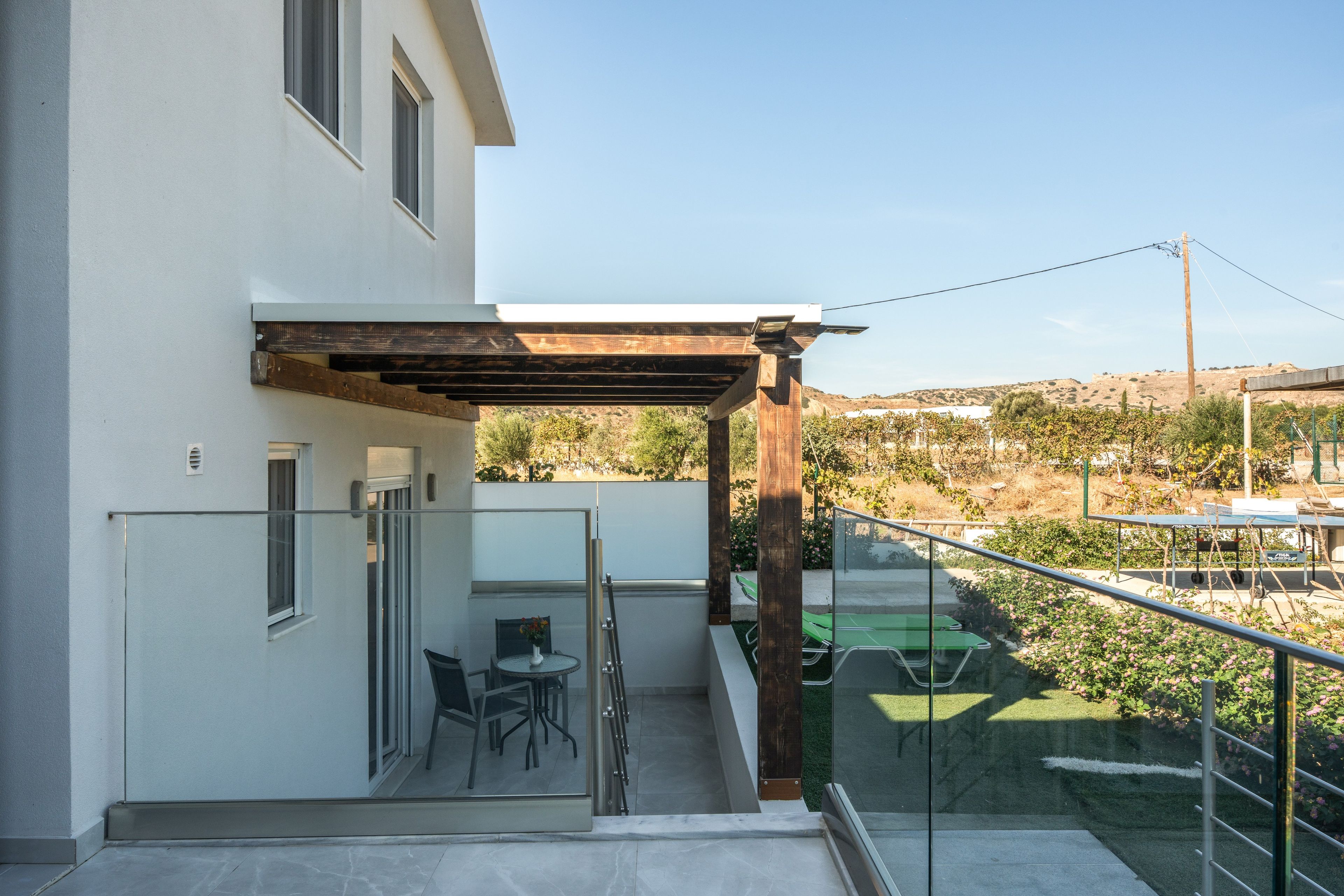 A modern building with a covered outdoor seating area, a glass door entrance, and a view of a hilly landscape.