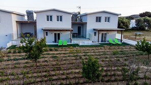 A modern two-story house with a flat roof, a covered porch, and a small garden area with green chairs.