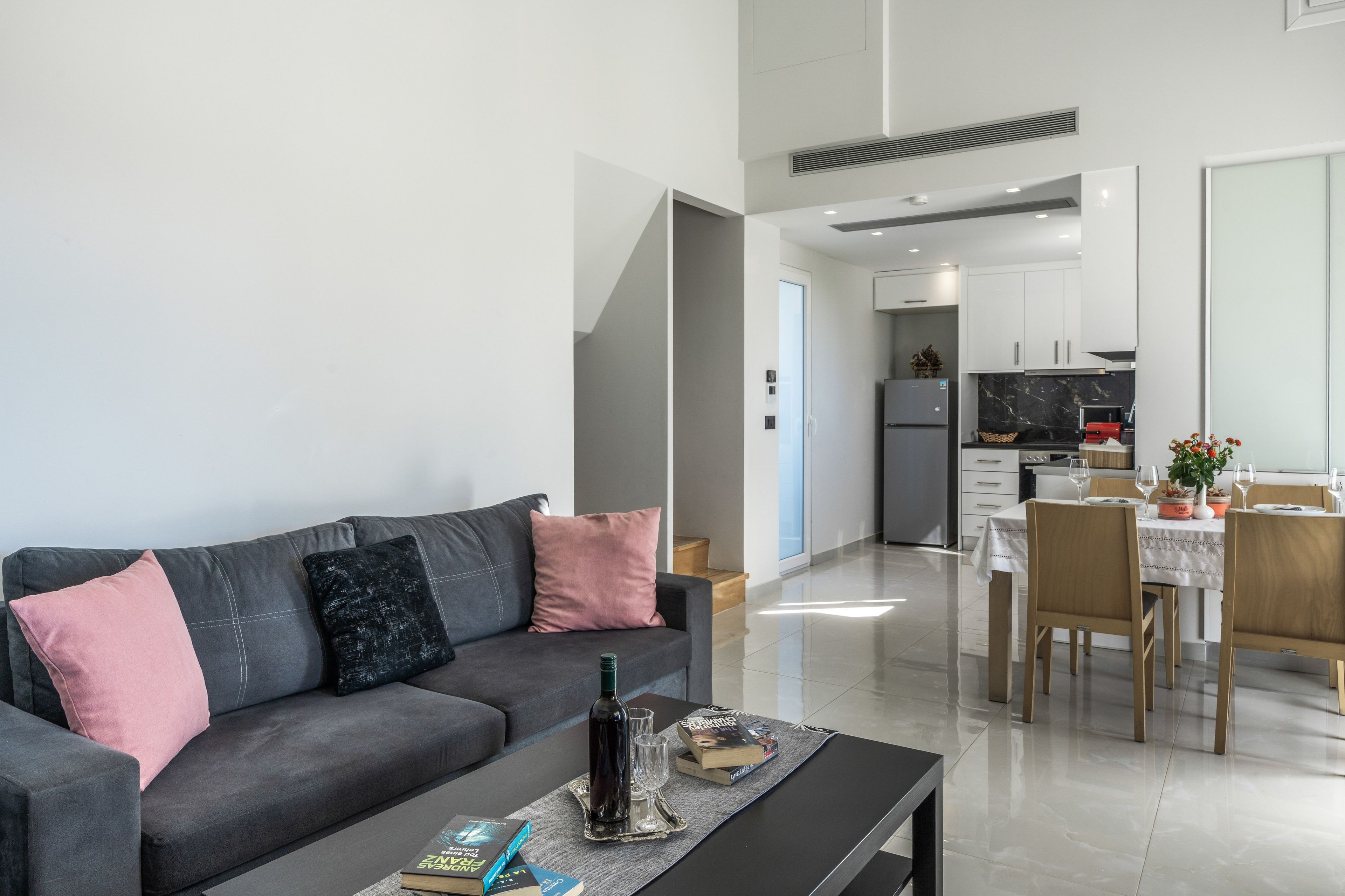 A modern living room with a grey sofa, a coffee table with books and a bottle, and a dining area with a table set for two.