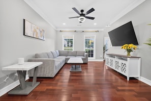 A modern living room with a grey sectional sofa, a white coffee table, a TV on a white cabinet, and a ceiling fan.