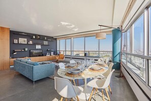 Ein modernes Wohnzimmer mit großem Fenster, einer Essecke mit rundem Tisch und einem blauen Sofa.