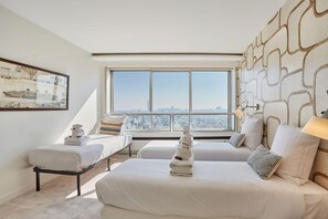 Ein Hotelzimmer mit großem Fenster, einem Bett mit weißen Bettwäsche, einem Nachttisch und einem an der Wand hängenden Kunstwerk.