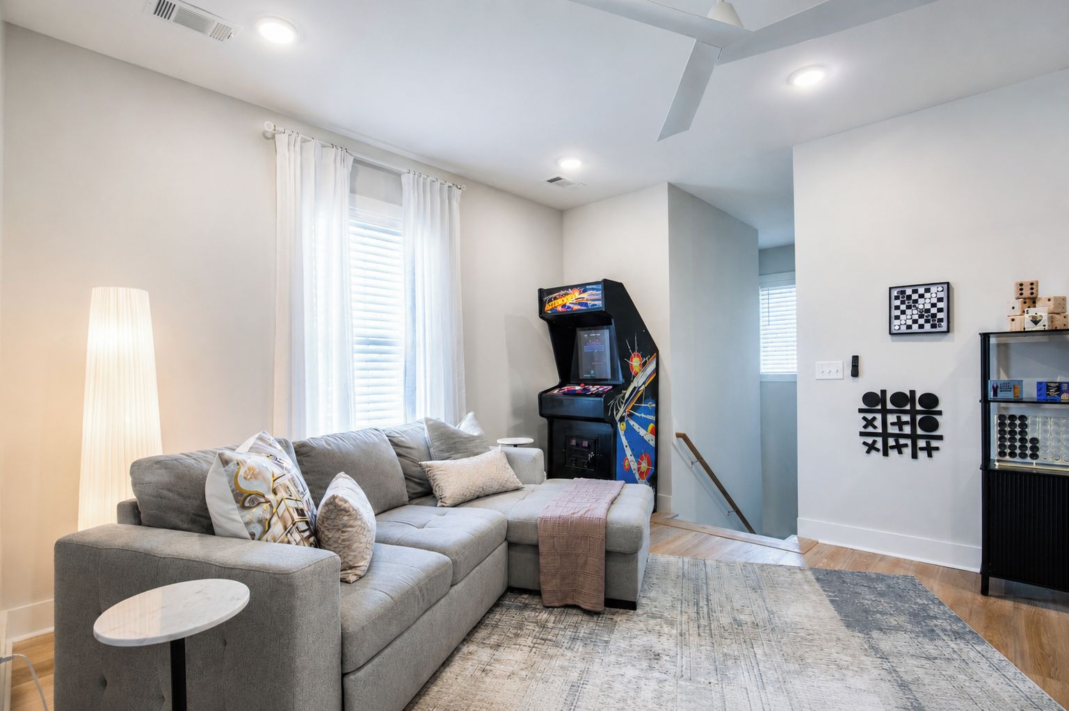 A modern living room with a grey sofa, a small round table, a gaming console, and a chess set on the wall.