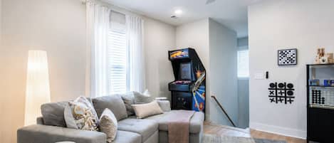 A modern living room with a grey sofa, a small round table, a gaming console, and a chess set on the wall.