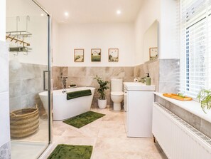 A modern bathroom with a freestanding bathtub, a toilet, and a sink. There are three framed pictures on the wall, a potted plant, and a window with blinds. - The Swallows (Lancaster)