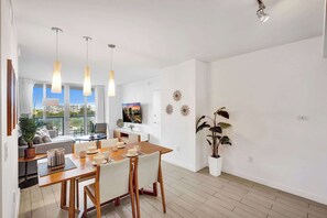 A modern dining area with a wooden table, chairs, and pendant lights. There is a sofa and a TV in the background. - Cozy Stunning 1-BR APT - Steps from the Ocean (Hallandale Beach)