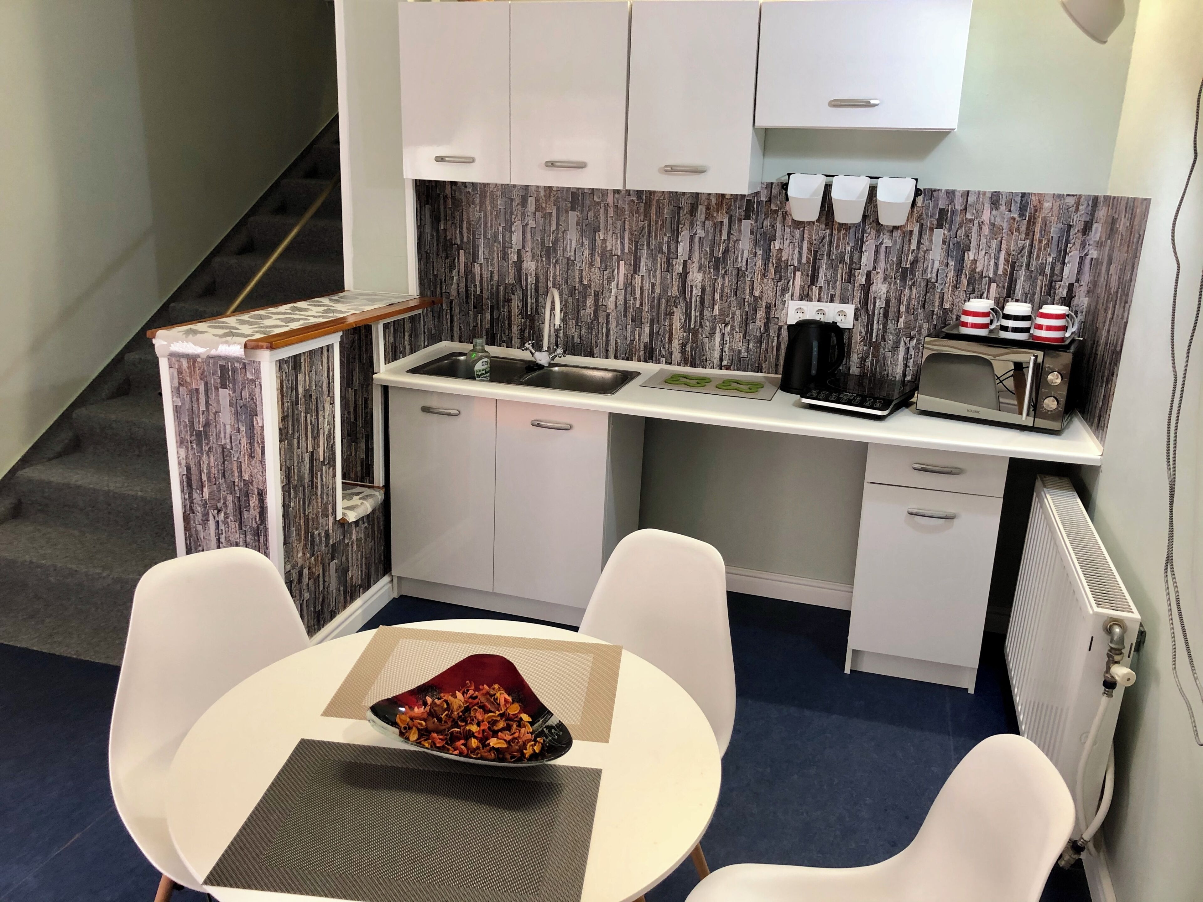 A compact kitchenette with a sink, a coffee maker, and a toaster. There is a small table with a bowl of snacks and two white chairs.