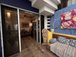 A room with a sliding glass door leading to a dining area, a Buddha statue, a sofa with patterned cushions, and a large abstract painting on the wall.