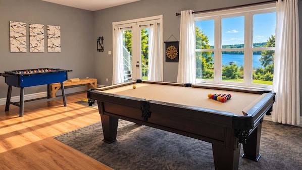 A pool table with a billiard cue and balls, a foosball table, and a dartboard in a room with a view of a lake.