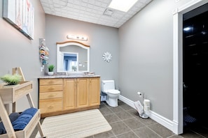 A modern bathroom with a wooden vanity, a white toilet, a large mirror, and a decorative wall piece.