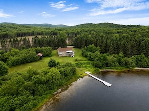 A house with a large lawn and a dock extending into a lake, surrounded by dense forest and hills.