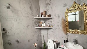 Ein Badezimmer mit Marmorwand, Toilette in Weiß, Waschbecken mit goldener Spiegelrahmung und Ablagefläche für Pflegeprodukte.