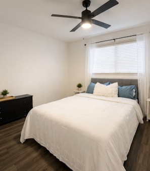 A bedroom with a bed, a ceiling fan, a nightstand, a dresser, and a window with blinds.