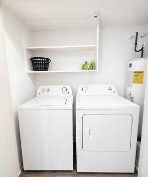 A laundry room with two white front-loading washing machines, a black laundry basket on a shelf, and a GE refrigerator with an ENERGY GUIDE sticker.