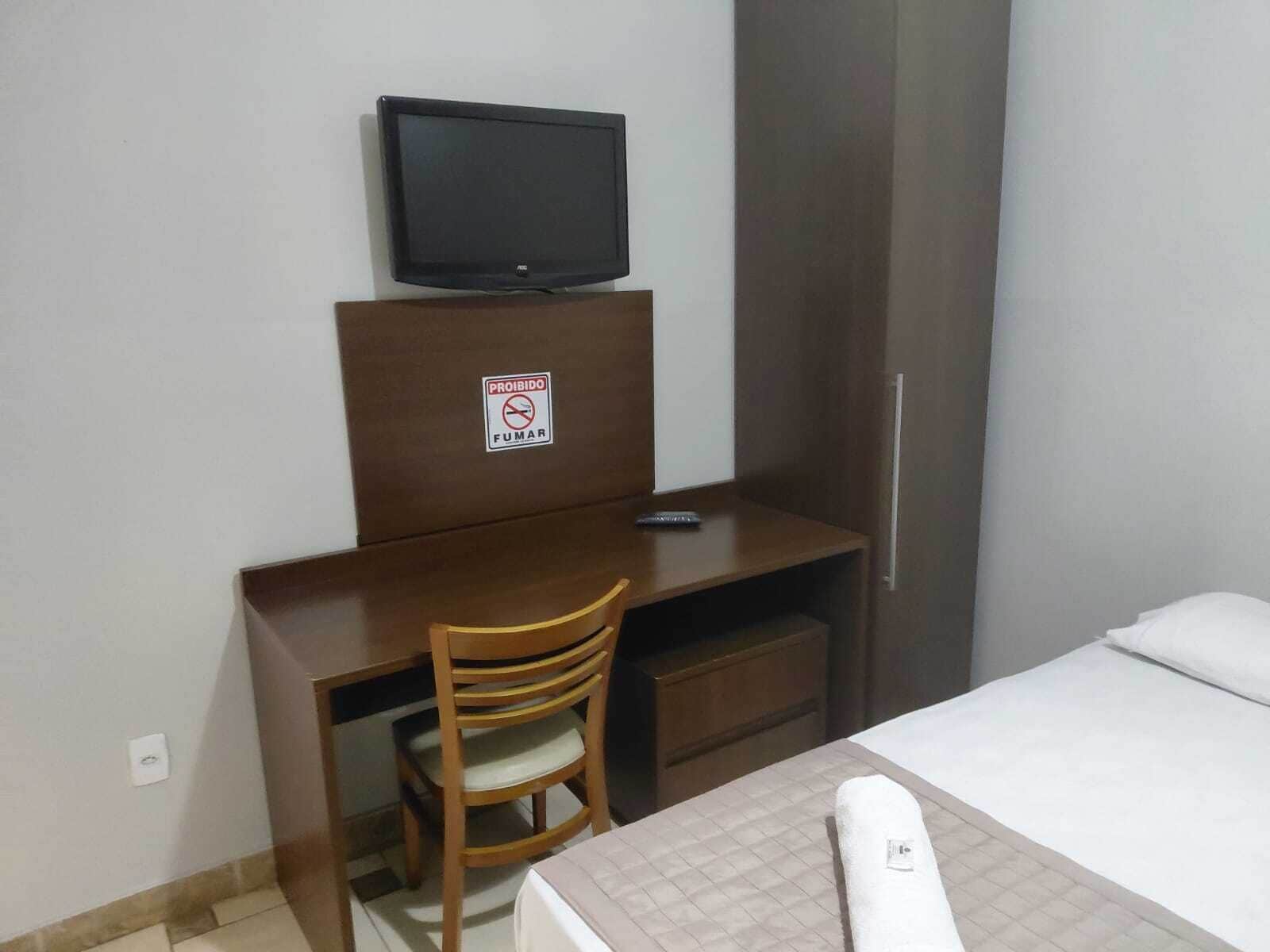 Quarto de hotel com cama, escrivaninha com televisão, cadeira e placa proibindo fumar.