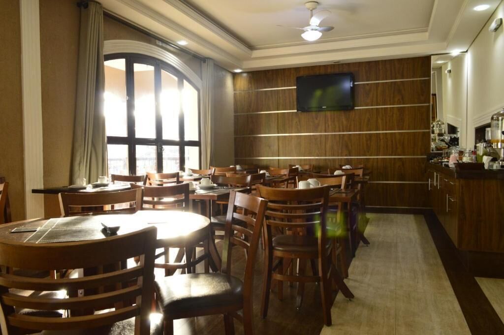 Uma área de refeições de hotel com mesas e cadeiras de madeira, uma janela grande com cortinas, uma televisão presa na parede e um espaço de cozinha com armários e eletrodomésticos.