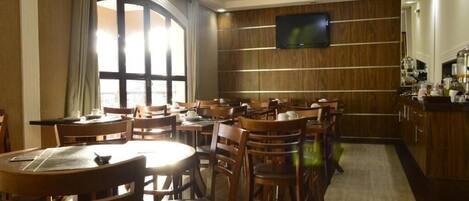 Uma área de refeições de hotel com mesas e cadeiras de madeira, uma janela grande com cortinas, uma televisão presa na parede e um espaço de cozinha com armários e eletrodomésticos.