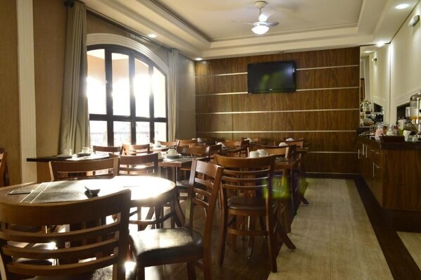 A hotel dining area with wooden tables and chairs, a large window with curtains, a television mounted on the wall, and a kitchen area with cabinets and appliances.