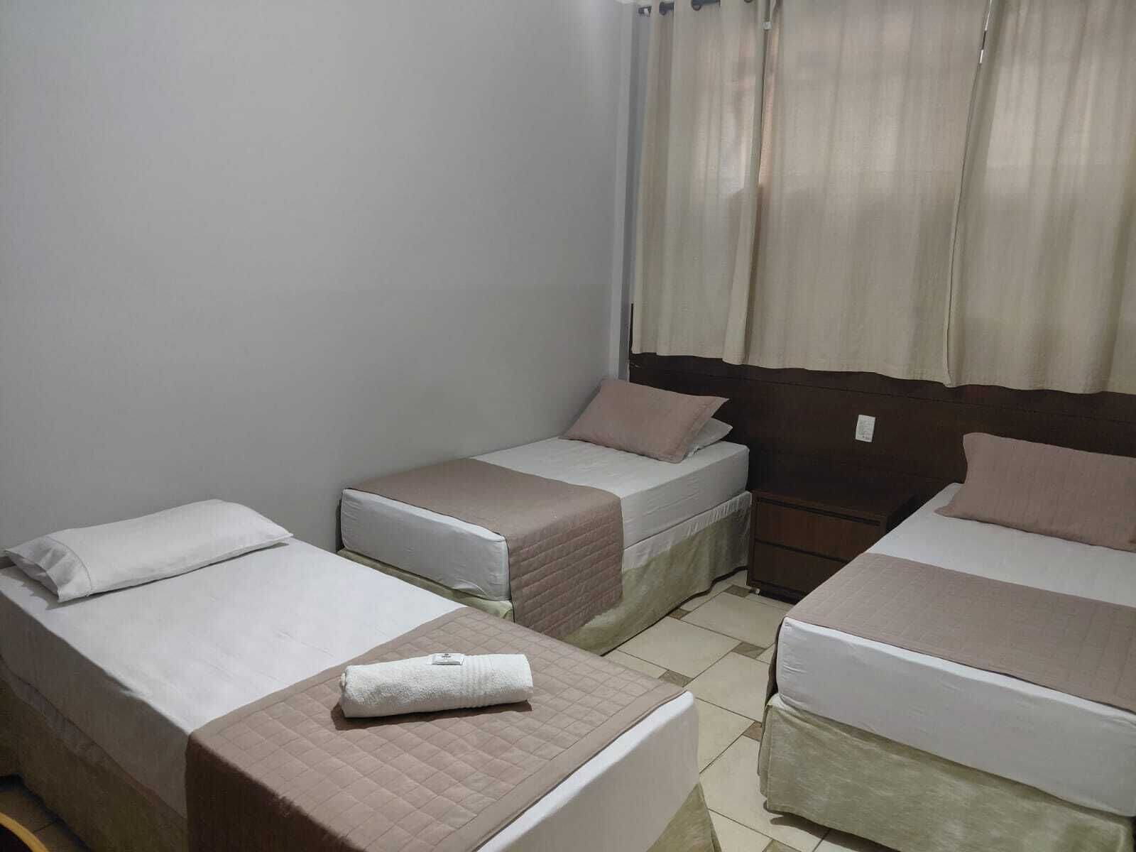Quarto de hotel com três camas de solteiro, uma pequena mesa de cabeceira e uma janela com cortinas.