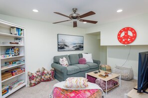 A living room with a sofa, a coffee table, a bookshelf, a ceiling fan, and a wall-mounted peace sign. - BeachRentalCarlsbad - 180° Ocean Views - 5 Min Walk to the Beach - Sleeps 7 (Carlsbad)