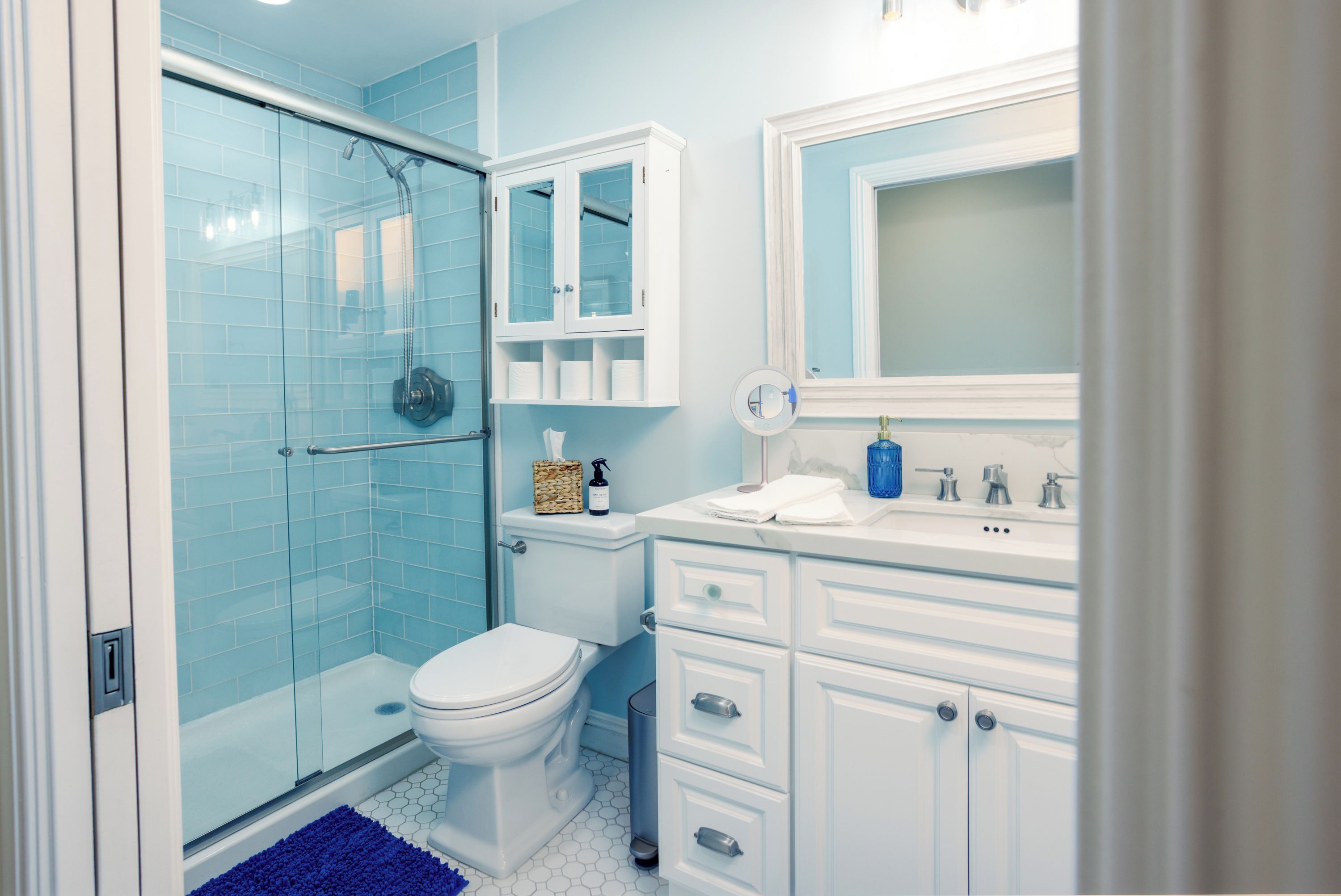 A bathroom with a glass shower enclosure, a toilet, a sink with a mirror, and a cabinet with toiletries.