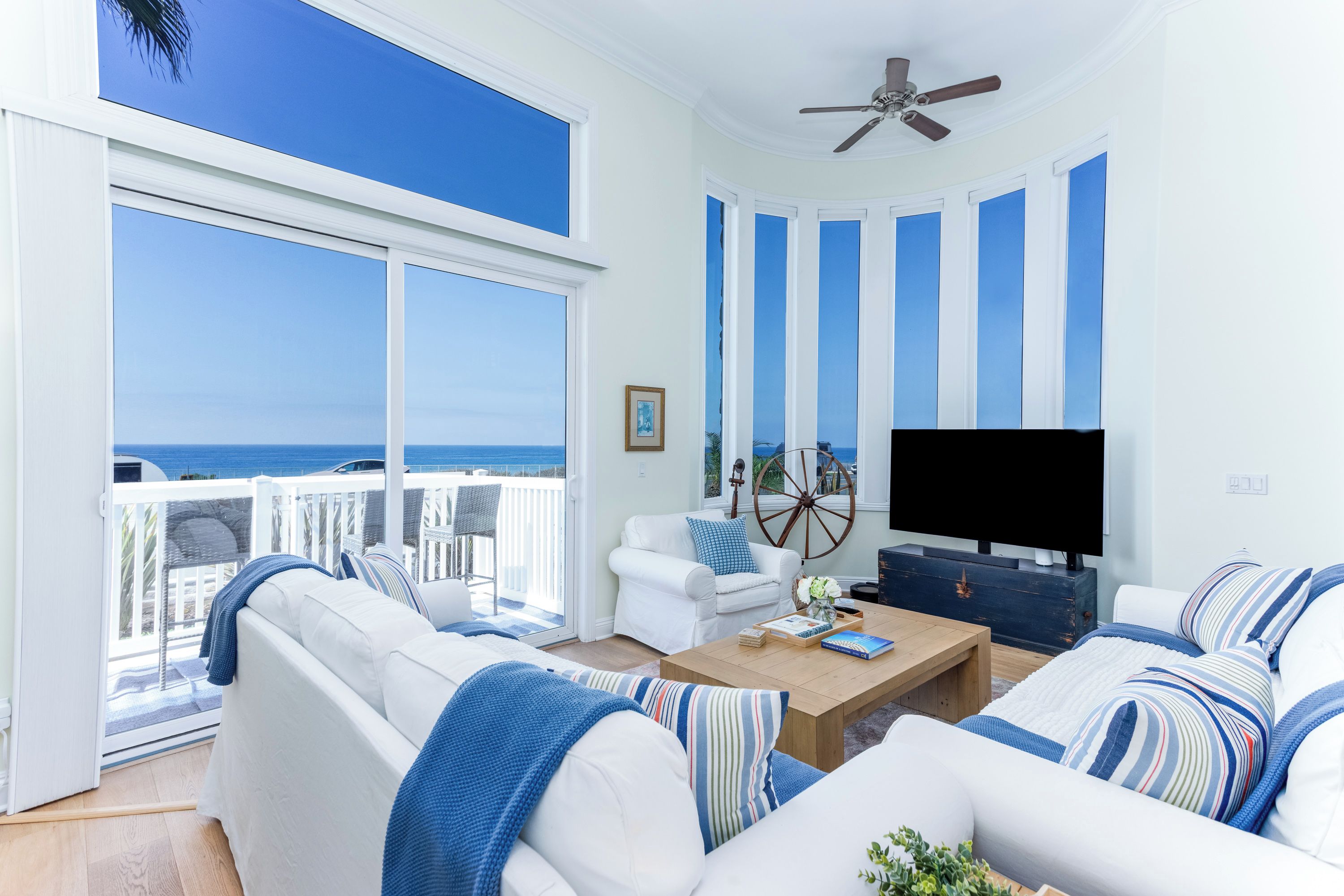A living room with a large window overlooking the ocean, a ceiling fan, and a television on a wooden stand.