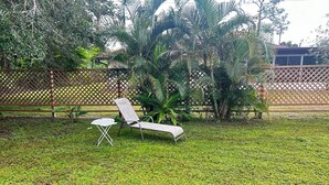 En gräsyta med en vit, hopfällbar stol och ett litet bord, omgiven av tät vegetation och ett trästaket.
