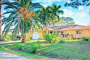 Une maison de plain-pied avec un jardin devant, des palmiers et un ciel dégagé.