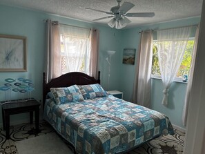 Un dormitorio con cama, mesita de noche, ventilador de techo y ventana con cortinas.