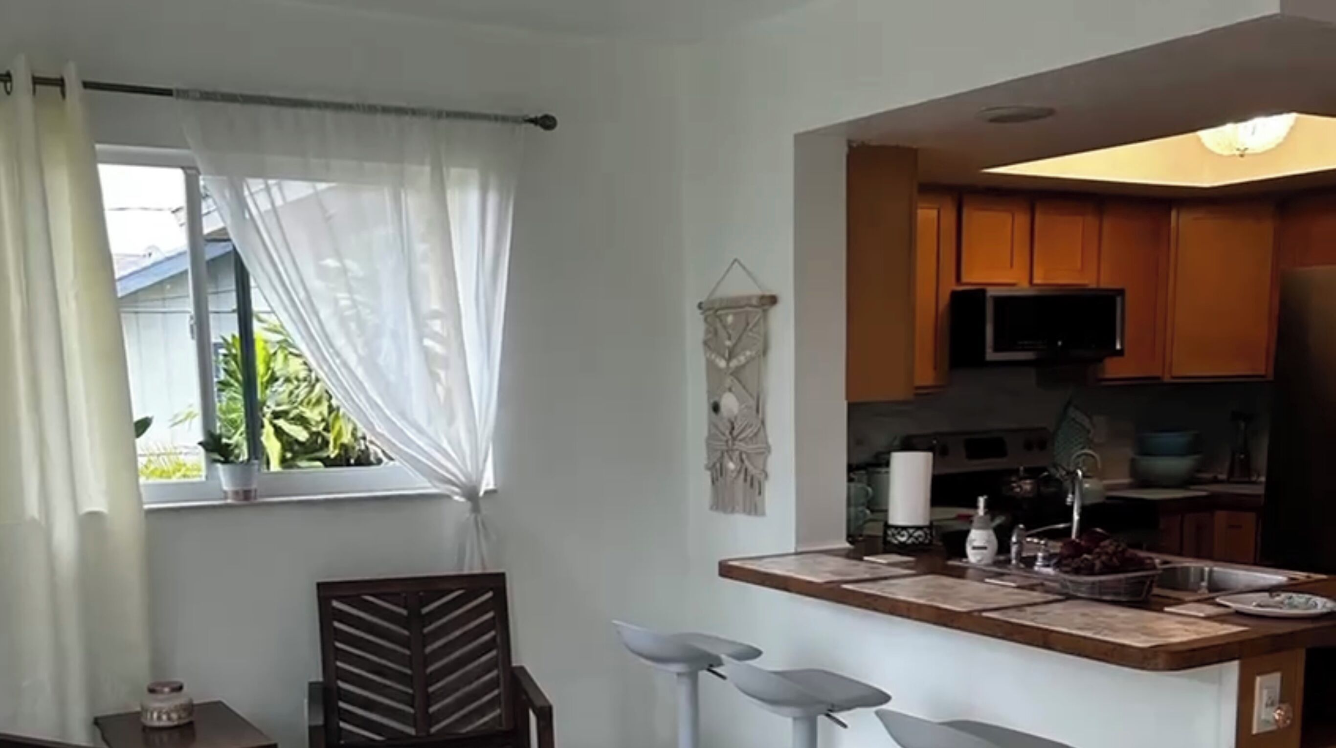 A modern kitchen with wooden cabinets, a microwave, and a sink. There is a window with sheer curtains and a decorative wall hanging.