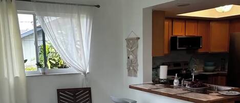 Una cucina moderna con mobili in legno, un forno a microonde e un lavandino. Si vede una finestra con tende leggere e un decoro a parete.