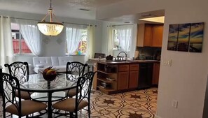 Un comedor con una mesa redonda y sillas, una araña, y vista a la cocina con armarios de madera y piso de baldosas.