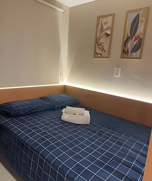 Ein kleines, ordentlich gemachtes Bett mit einer blauen, gemusterten Tagesdecke und Kissen, ein weißer Nachttisch und zwei gerahmte Kunstwerke an der Wand.