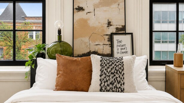 A bedroom with a bed, a green vase, a framed inspirational quote, and a view of buildings through large windows. - The Marle  (Baltimore)