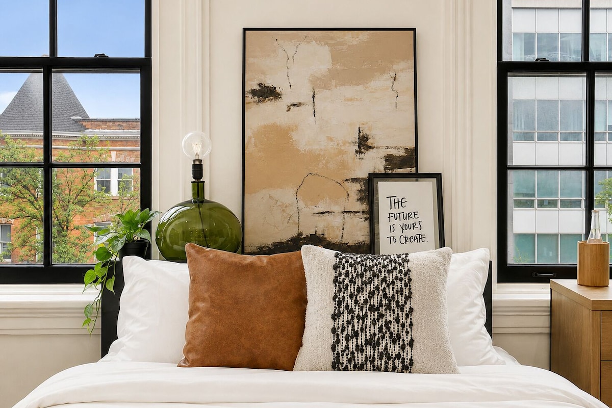 A bedroom with a bed, a green vase, a framed inspirational quote, and a view of buildings through large windows.