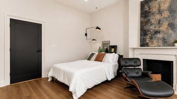 Un dormitorio con una cama, una silla, una chimenea y un cuadro en la pared.