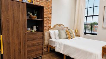 Un dormitorio con un armario de madera, una cama con ropa de cama blanca, una ventana con vistas y una pared de ladrillo.