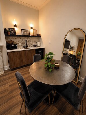 A dining area with a round table, four chairs, a potted plant, and a kitchen in the background.
