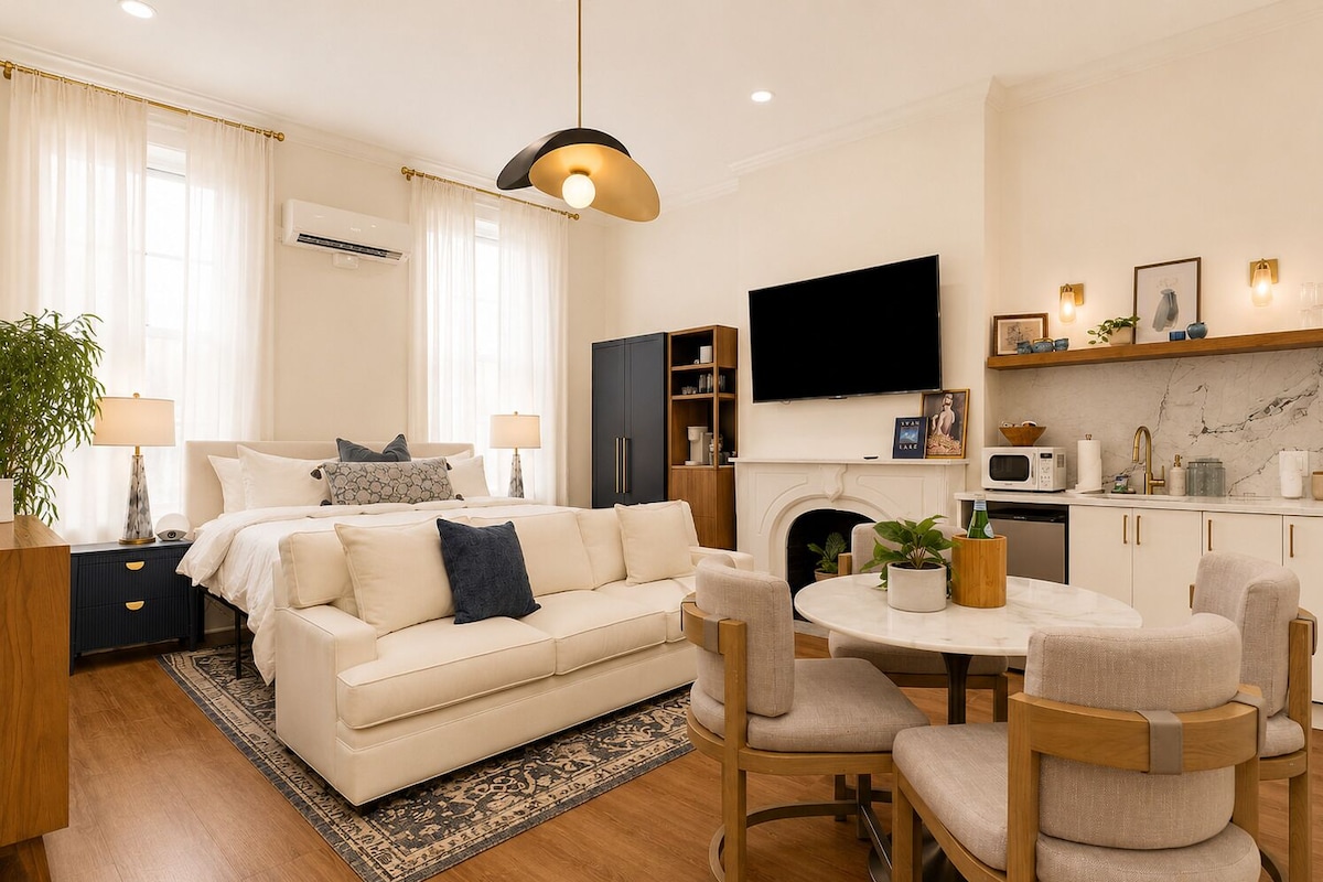 A modern hotel room with a bed, bedside tables, a desk, a dining area, and a kitchen.