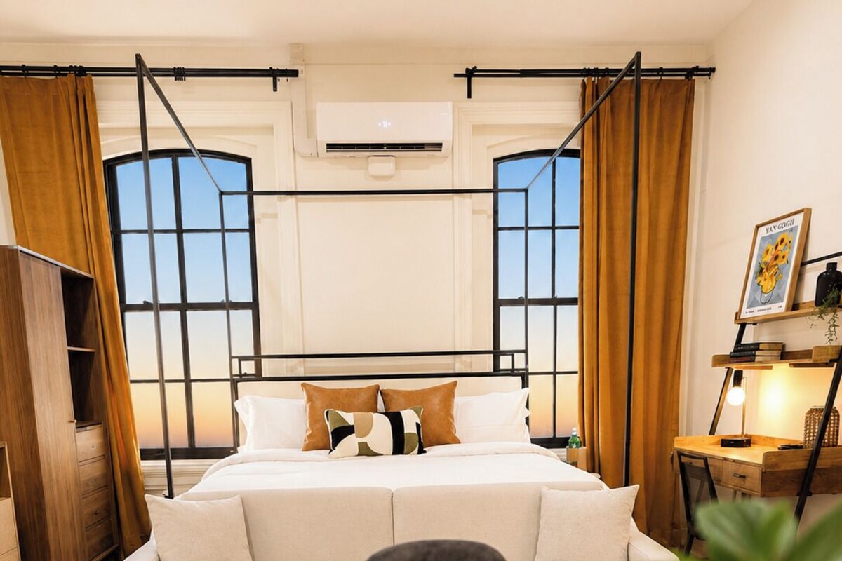 A bedroom with a bed, a sofa, a desk, a chair, a table, a plant, and a candle.