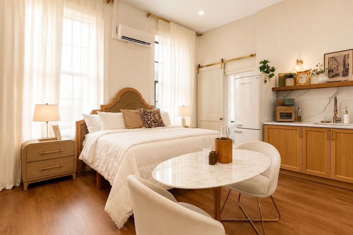 A hotel room with a bed, bedside tables, a round dining table, chairs, a kitchen area, and a window with curtains.
