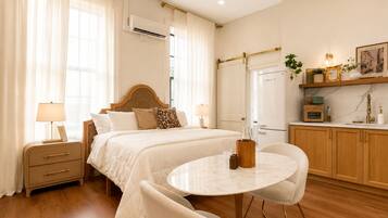 A hotel room with a bed, bedside tables, a round dining table, chairs, a kitchen area, and a window with curtains.