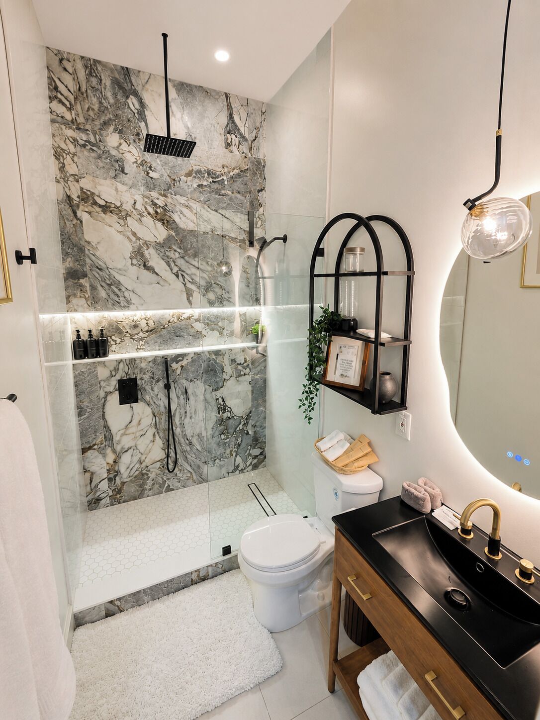 A modern bathroom with a marble wall, a glass-enclosed shower, a white toilet, and a black vanity with a gold faucet.