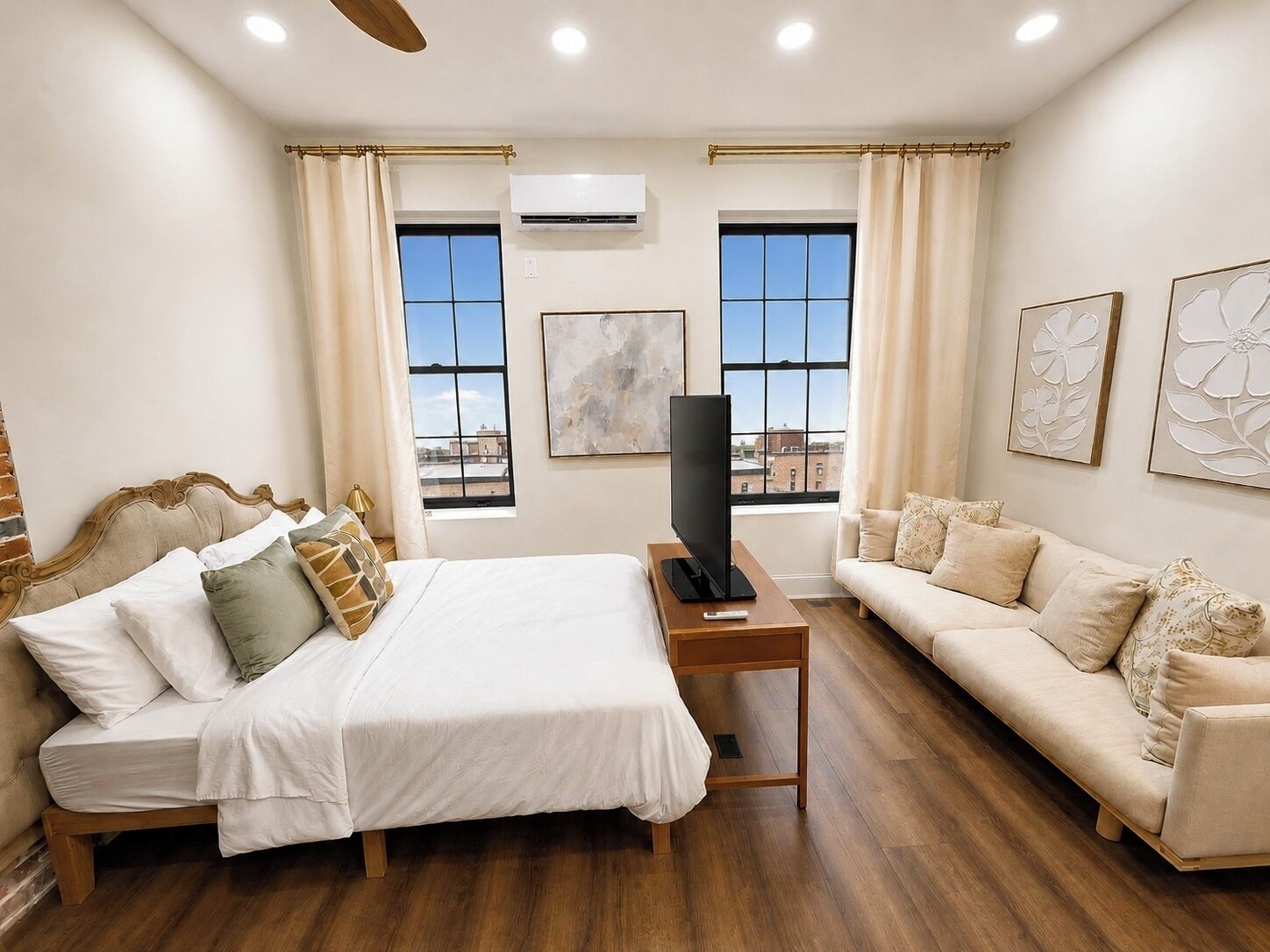 Habitación de hotel con una cama, un sofá, un televisor en un mueble bajo y dos ventanas con cortinas.