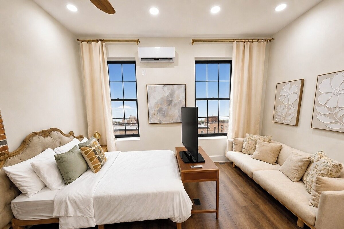 A hotel room with a bed, a sofa, a TV on a stand, and two windows with curtains.