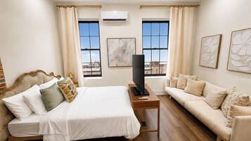 Habitación de hotel con una cama, un sofá, un televisor en un mueble bajo y dos ventanas con cortinas.